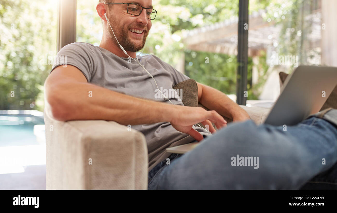 Smiling mature man using his laptop at home, he is sitting on couch with earphones. Stock Photo