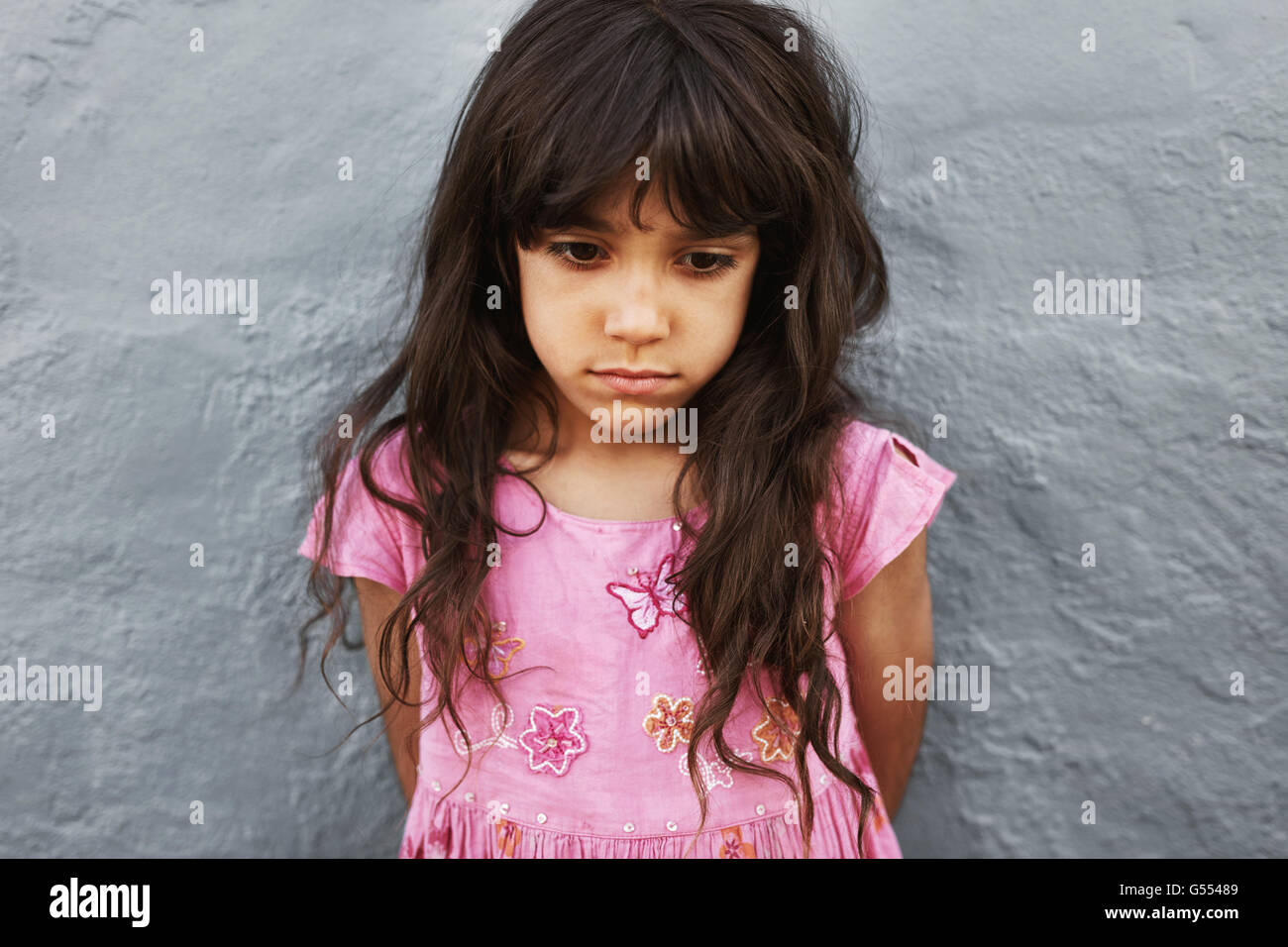 Close up portrait of little girl standing looking upset. Sad young girl standing against a grey wall. Stock Photo