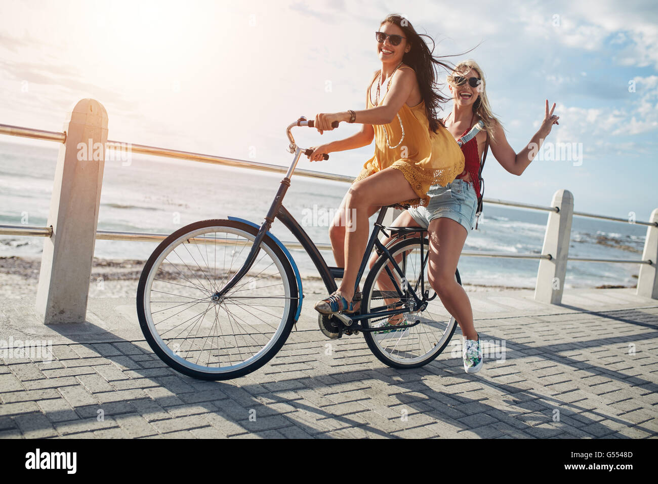 Two stylish young female friends on a bicycle along seaside. Best friends enjoying a day on bike. Stock Photo