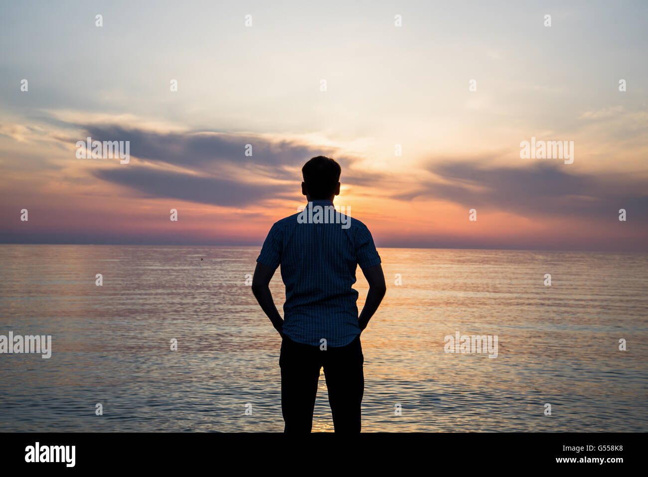 Young man standing at the beach in front of amazing sea view at sunset or sunrise and thinking about his future. Rear view Stock Photo