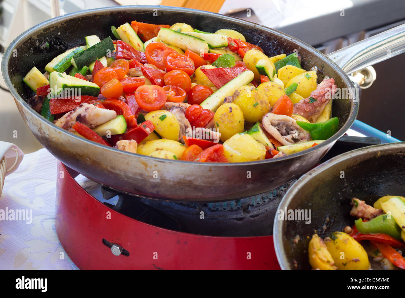 Vegetables fry on a large frying pan on fire. Cooking at the festival. Girl  chef pouring vegetables into disposable utensils. Stock Photo