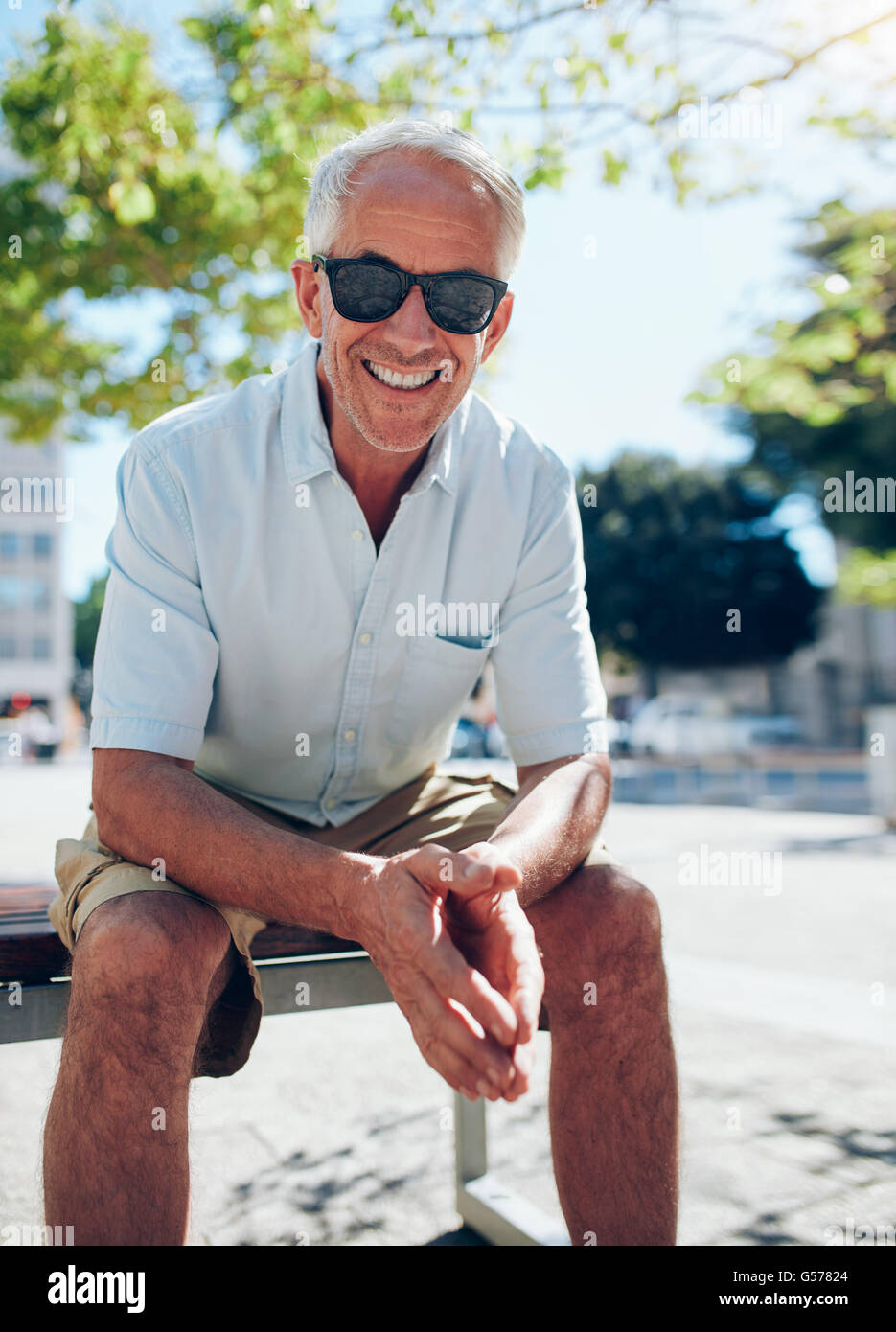 Portrait of smiling senior tourist sitting outdoors on a bench. Mature male tourist with sunglasses relaxing in the city on a su Stock Photo