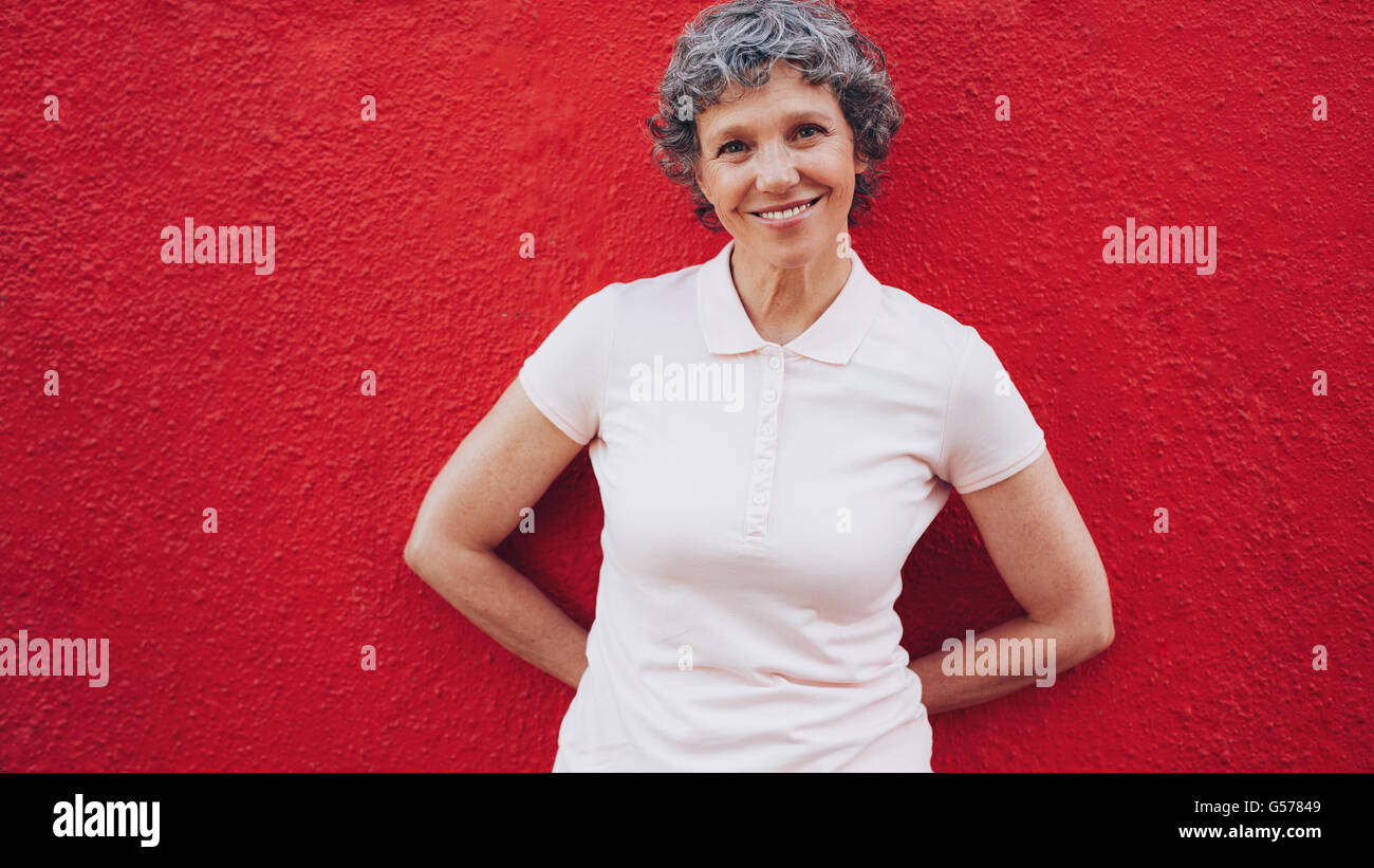 Portrait of happy middle aged woman standing relaxed against red background. Cheerful mature woman leaning to a red wall. Stock Photo
