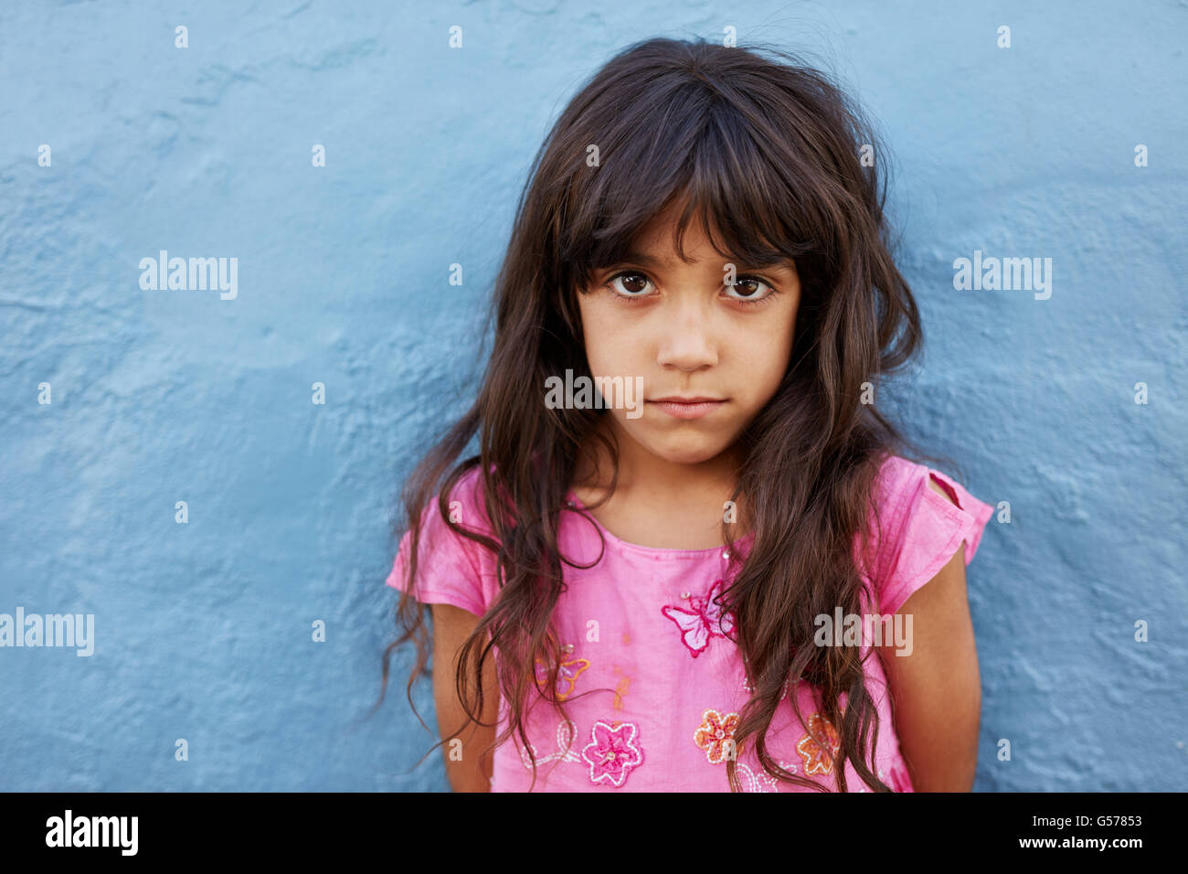 Close up portrait of innocent little girl standing against blue wall, she is looking at camera with serious expression on her fa Stock Photo
