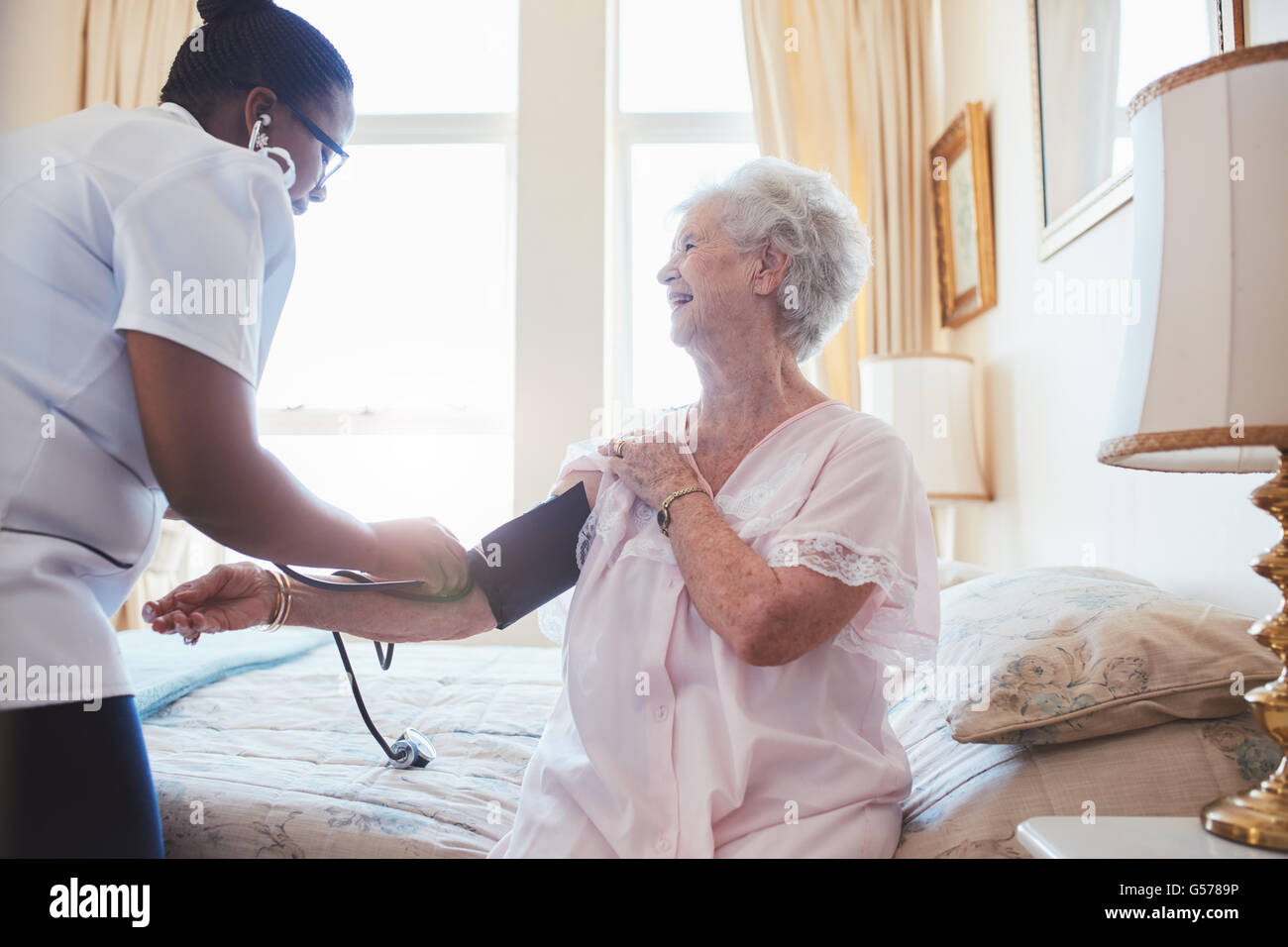 Nurse visiting senior female patient at home and taking blood pressure. Old woman sitting on bed. Stock Photo