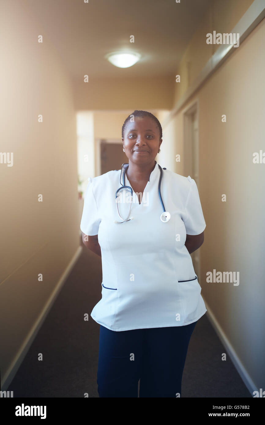 Portrait of happy african female nurse with stethoscope standing in corridor of a nursing home. Caring female doctor looking at Stock Photo