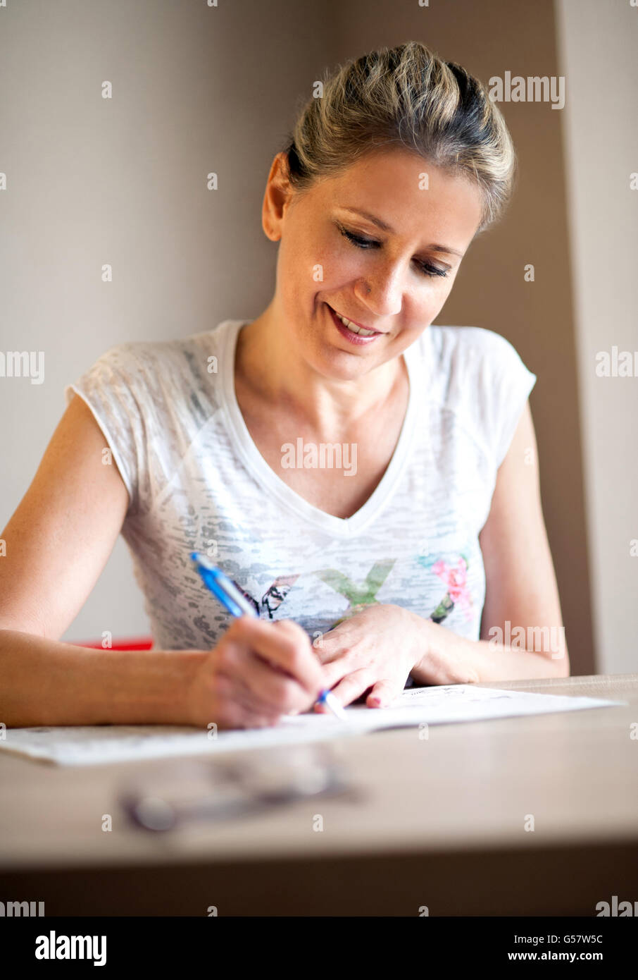 Single beautiful mature woman with pretty smile and tied back hair writing notes or filling out a form with pen at table indoors Stock Photo