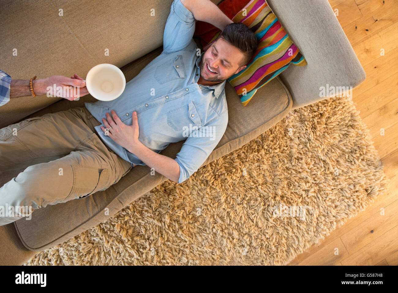 Man relaxing on the sofa. His partners arm is in the shot with an empty coffee cup in his hand as he takes it away. Stock Photo