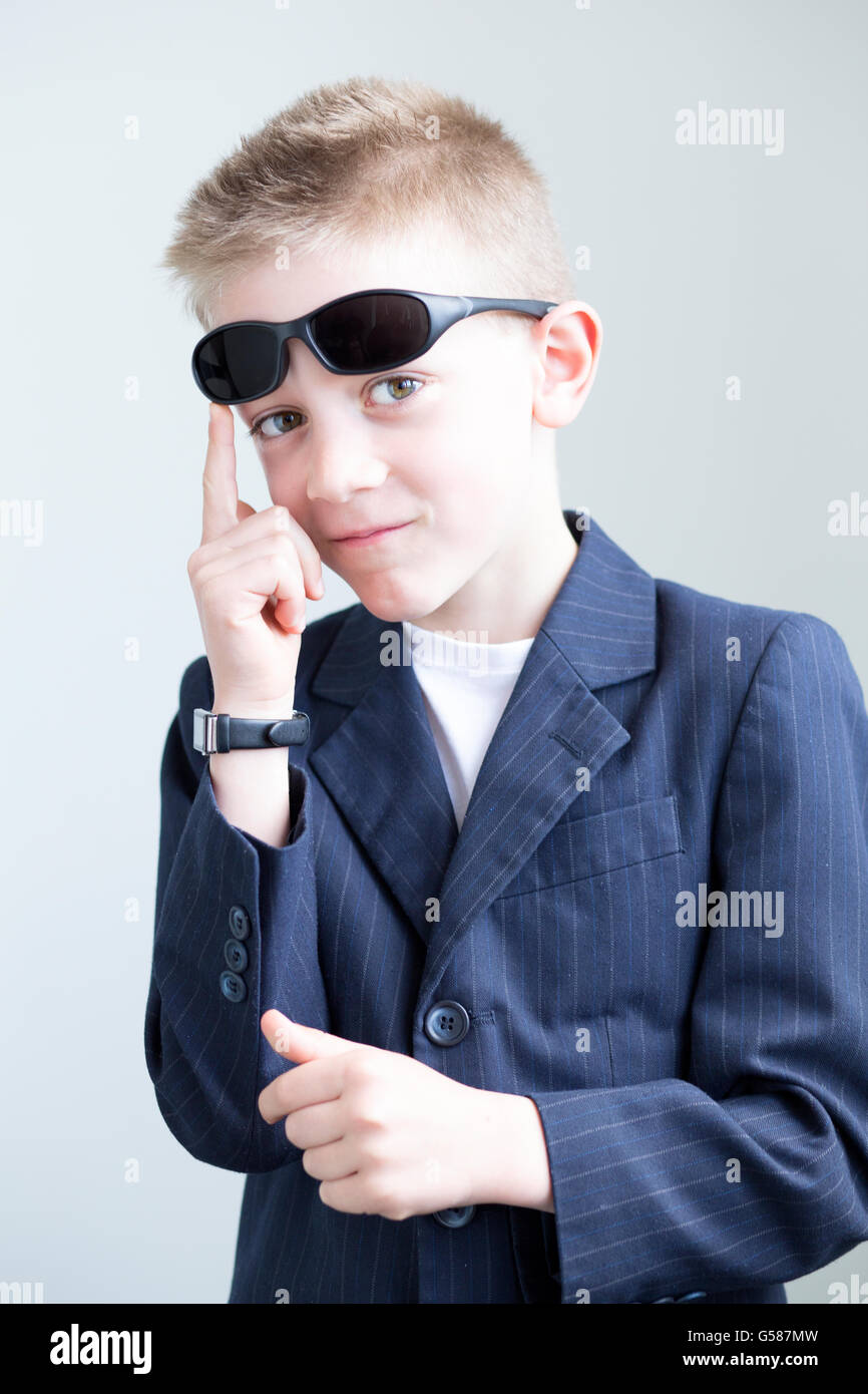 Young boy dressed up and posing like a spy. He is wearing a suit with sunglasses on his head and a formal watch. He is miming a Stock Photo