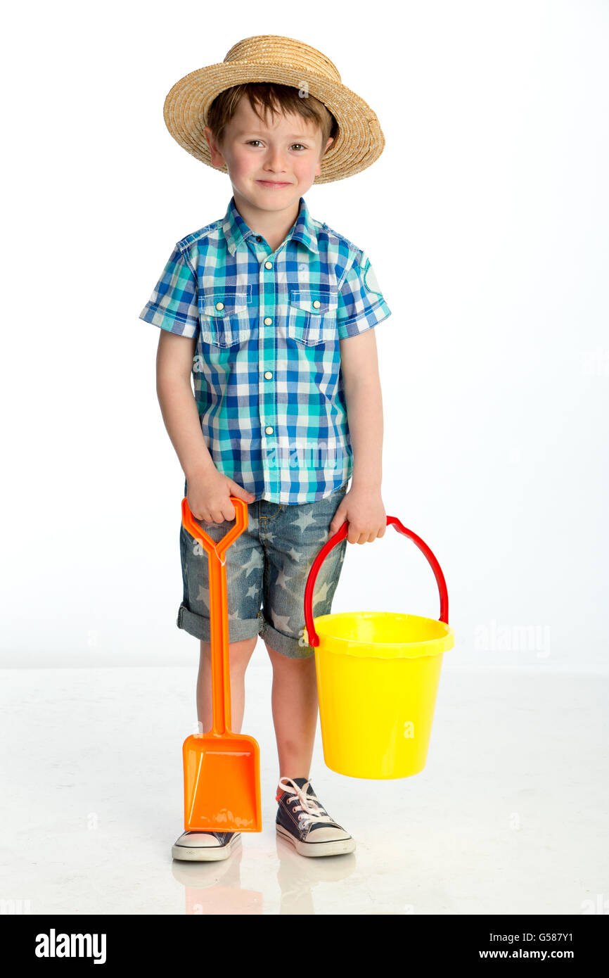 Cute little boy looking ready to go the beach. He is holding a bucket and spade and wearing a straw hat. He is smiling for the c Stock Photo