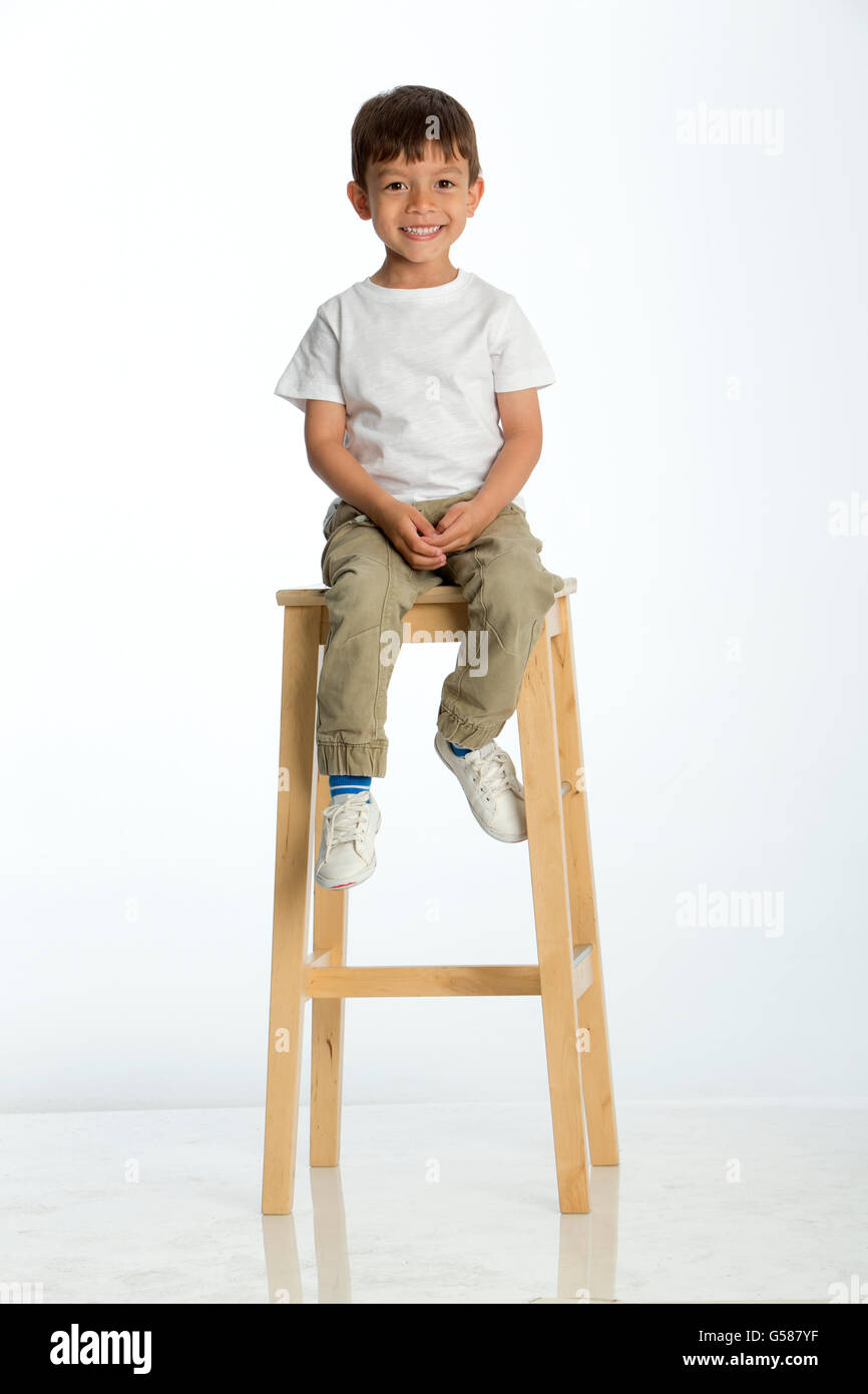 Cute young boy sitting on a stool against a white background, smiling for the camera. Stock Photo