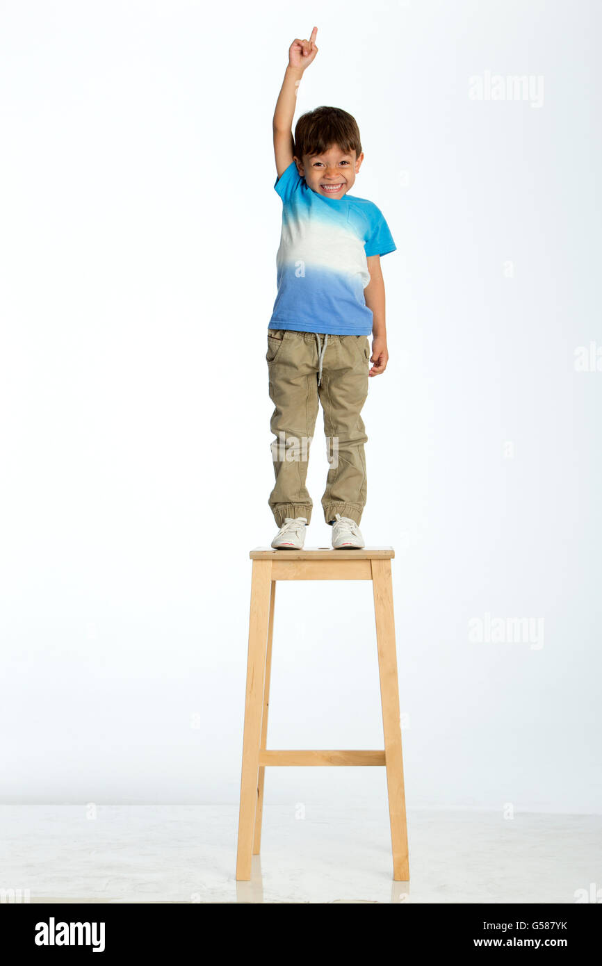 Little boy standing on a high stool, pointing up to the ceiling. He is against a white background. Stock Photo