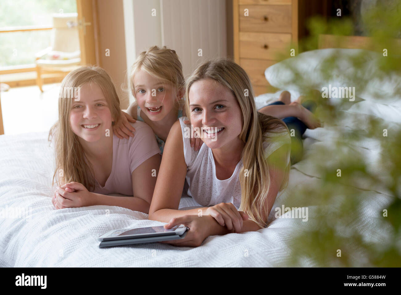 Sisters lying on a bed in their home with a digital tablet, smmiling for the camera. Stock Photo
