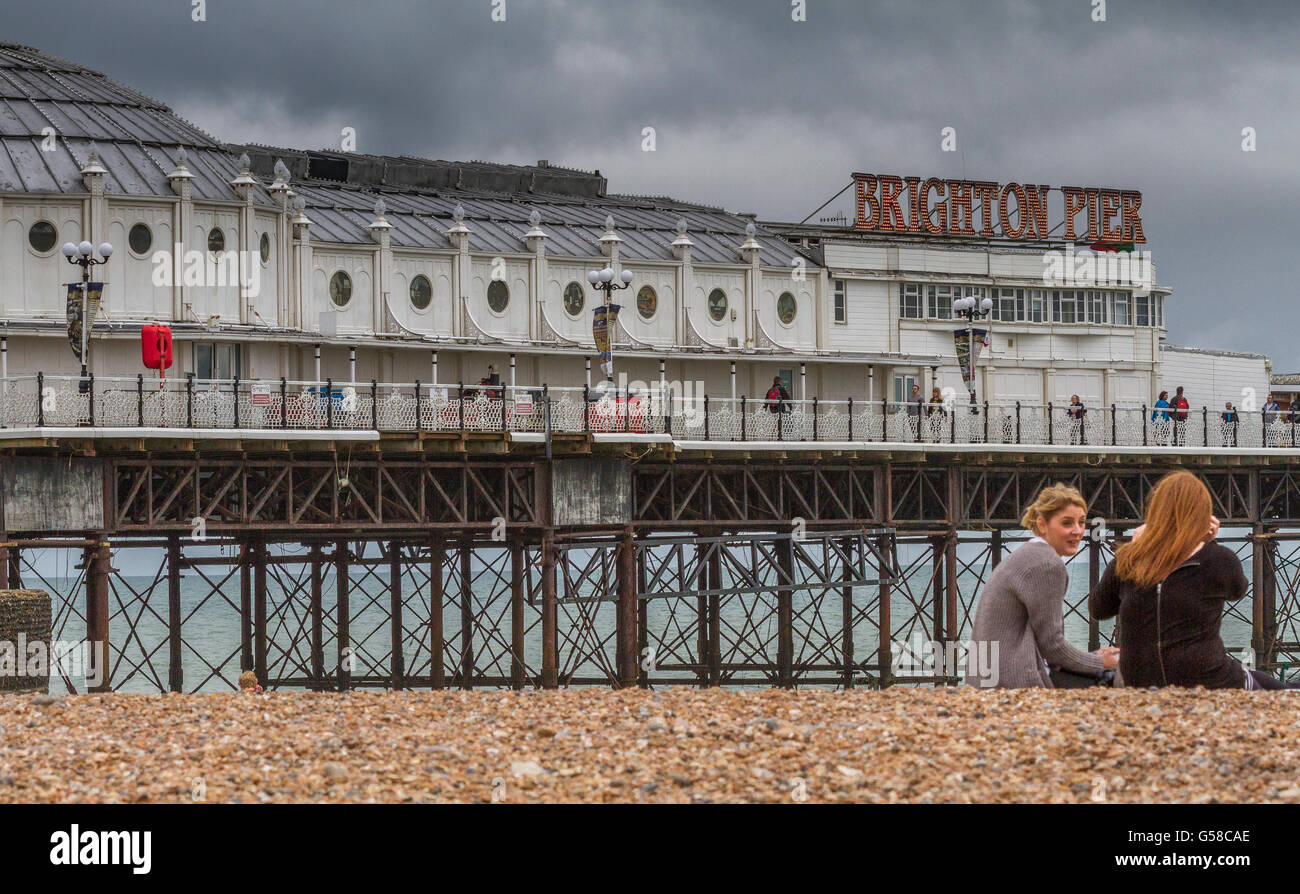 Two girls sitting on the pebbles of Brighton Beach talking in front of Brighton Palace Pier,Brighton , East Sussex , UK Stock Photo