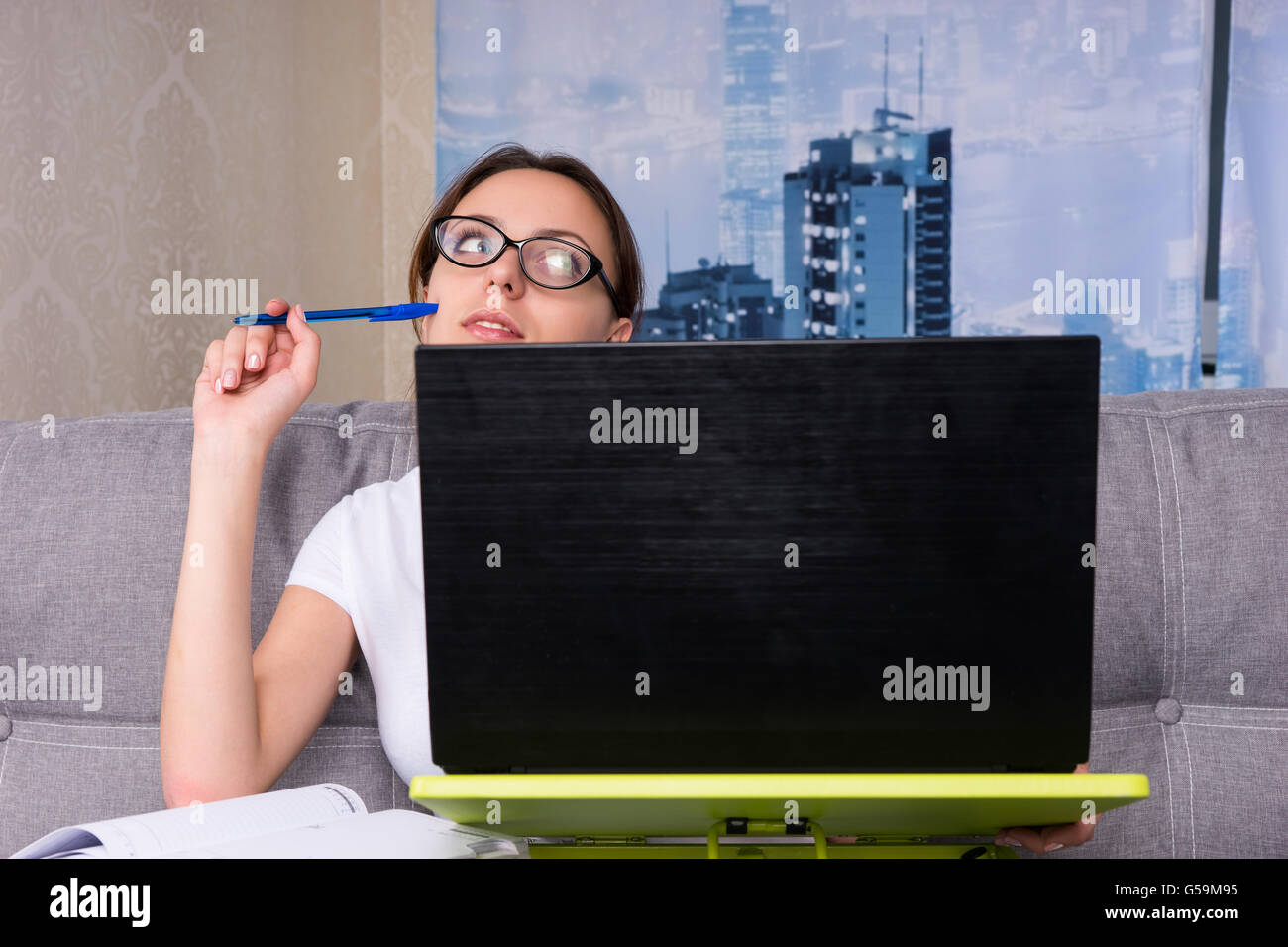 Dreamy young woman has lost in thoughts while working on a laptop and doing her business from home holding a pen sitting on a sofa. Stock Photo