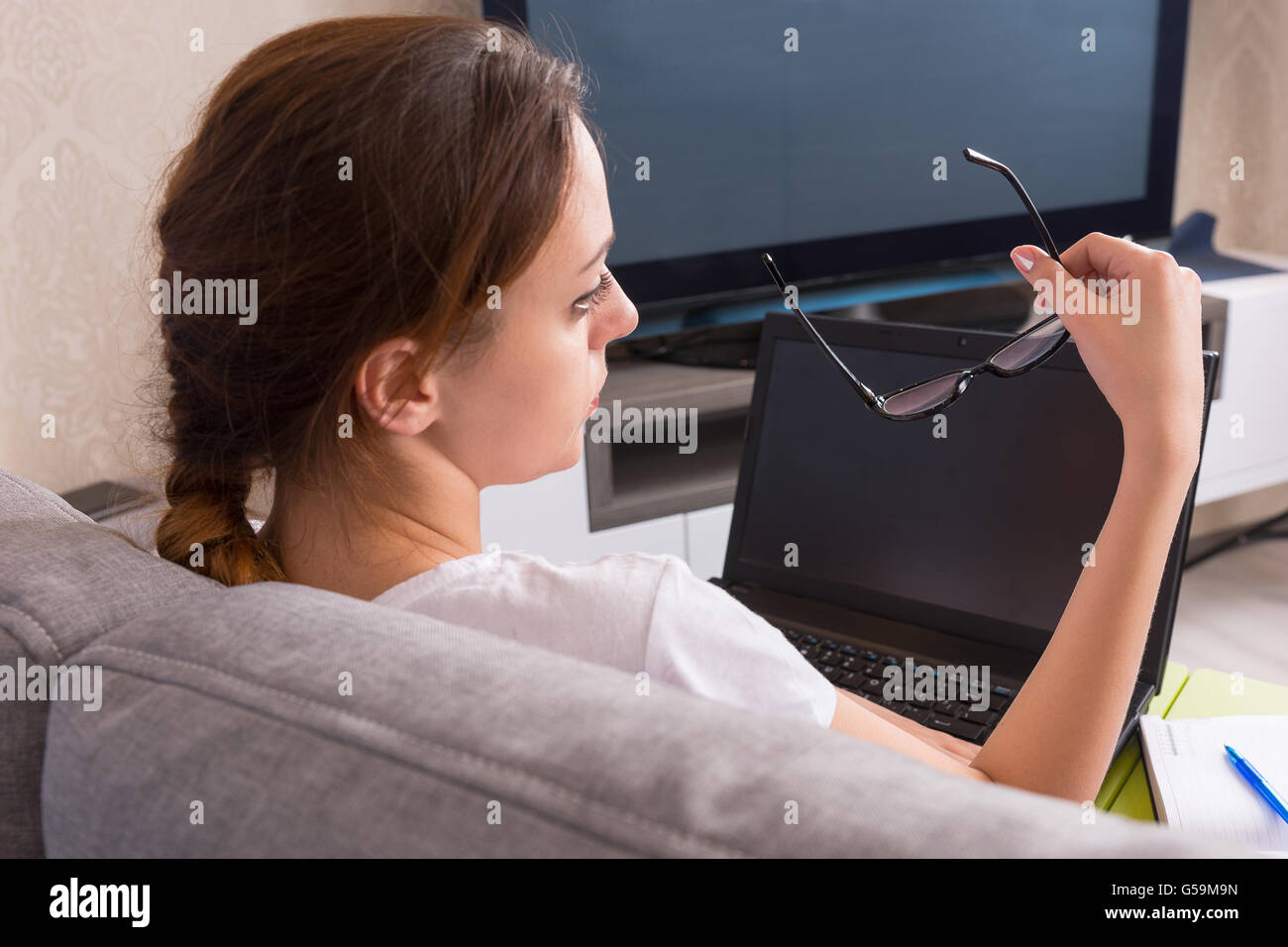 Back view of young woman holding glasses while working on a laptop doing her business from home sitting on a sofa in the living Stock Photo