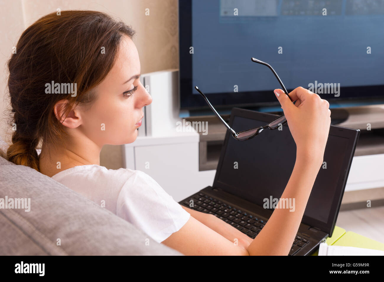 Back view of attractive woman holding glasses while working on a laptop doing her business from home sitting on a sofa in the living room  in a relaxed atmosphere. Stock Photo