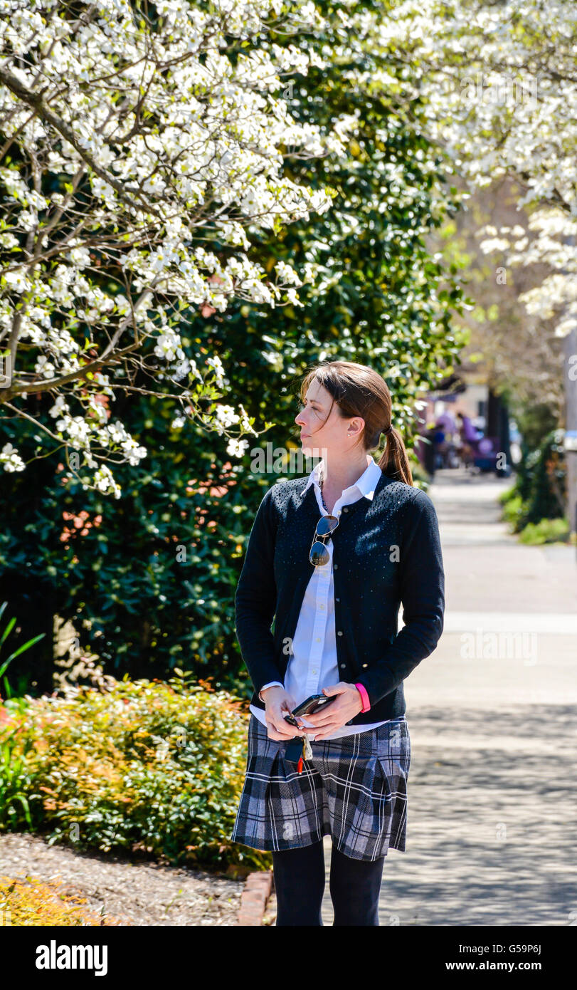 Profile young woman on sidewalk surrounded by blooming dogwood trees wearing parochial school style skirt & cardigan holding cell phone in USA Stock Photo