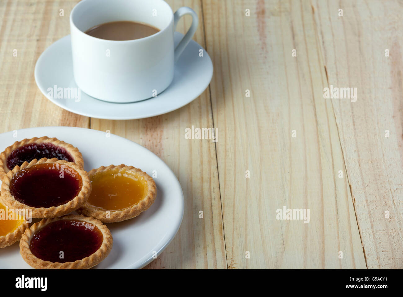 Plate of assorted fresh jam tarts on a wooden table with a cup of white coffee Stock Photo