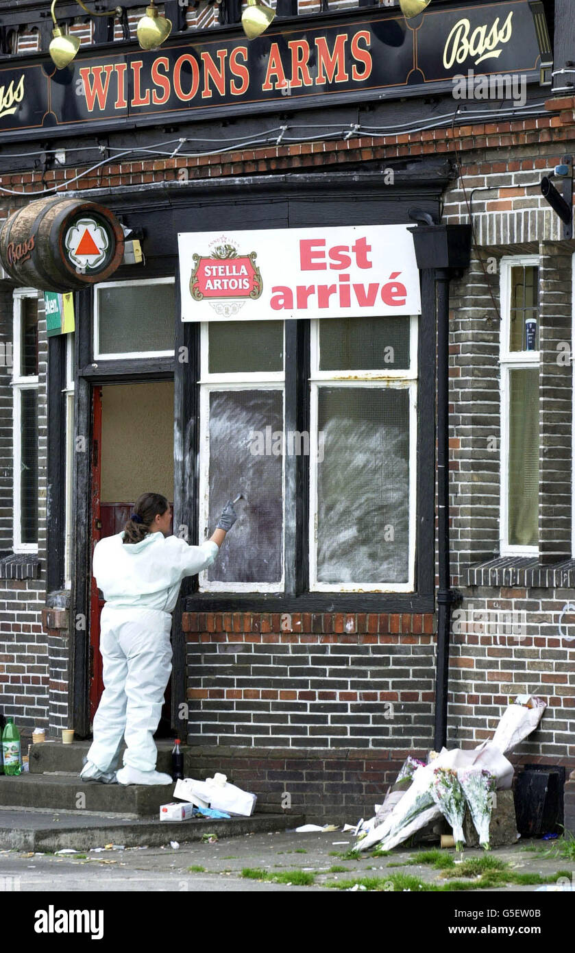 A police officer checks for fingerprints on windows at the Wilsons Arms Pub, Leeds, where a father-of-one was gunned down while children played party games. Former professional Rugby League star David Nelson, 38, was shot in the head at the pub in Seacroft on 22/07/01. *...Floral tributes to Mr Nelson are piled on the right. Stock Photo