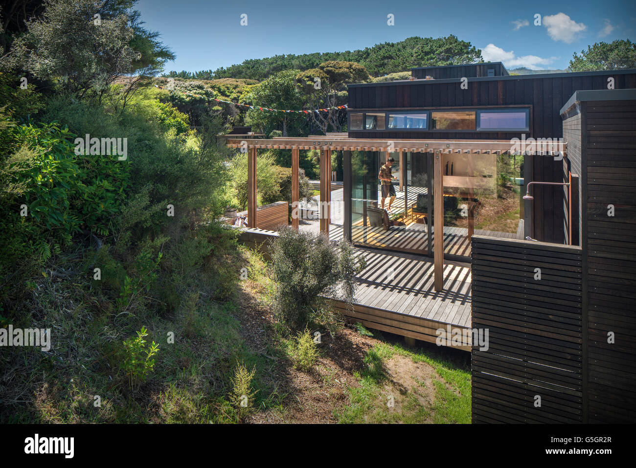 Side of house with wooden sun slats and decking and surrounding bush / countryside. Blue sky and sun. PEKA PEKA II HOUSE, Peka Peka - Kapiti Coast, New Zealand. Architect: HMA, 2016. Stock Photo