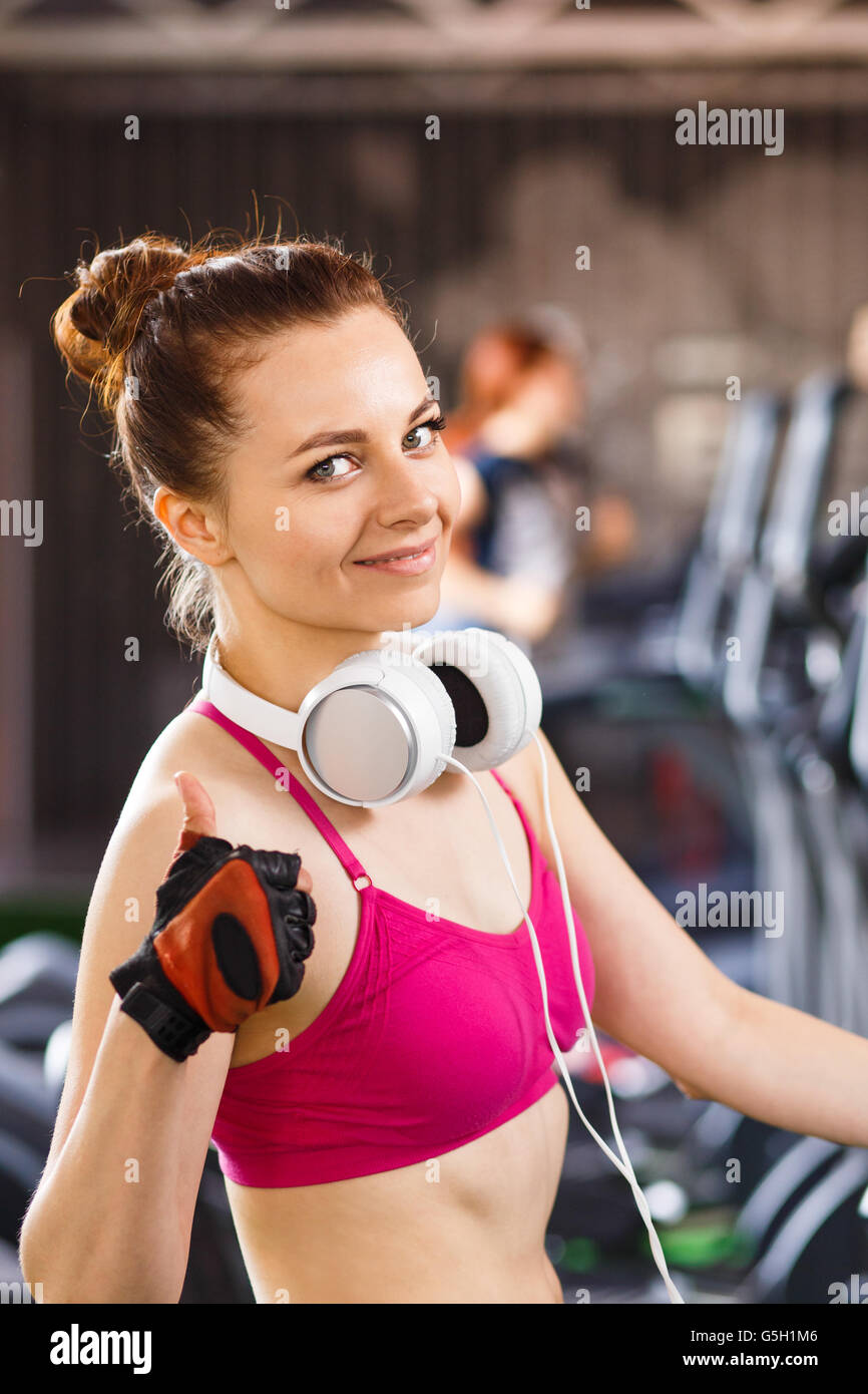 Young happy woman with headphones on cardio training in fitness center. Smiling beautiful girl showing thumbs up in cardio area  Stock Photo
