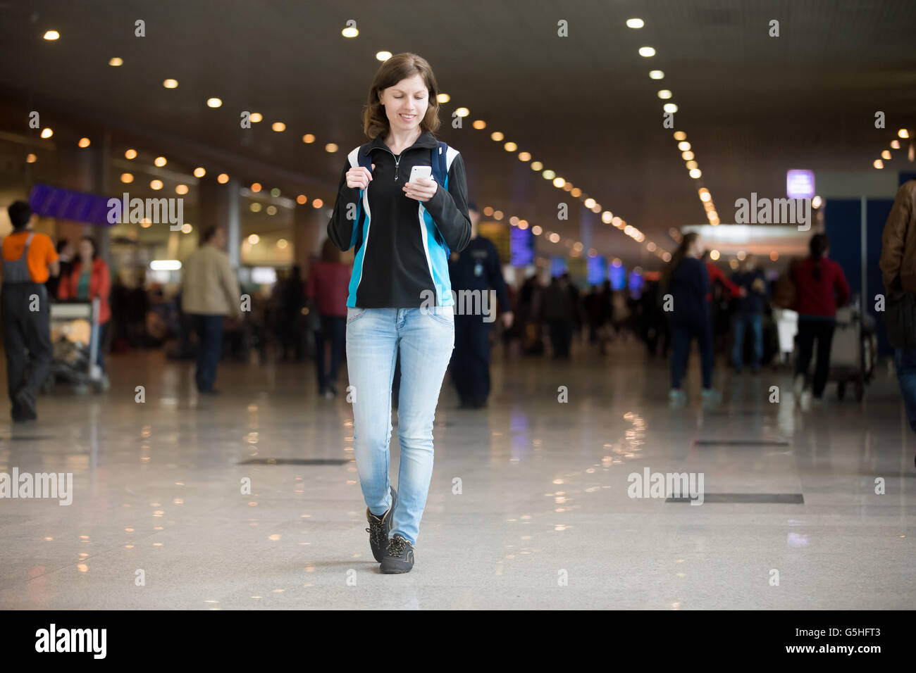 Smiling young woman in 20s with backpack walking in airport terminal, using cell phone app, messaging, wearing jersey Stock Photo