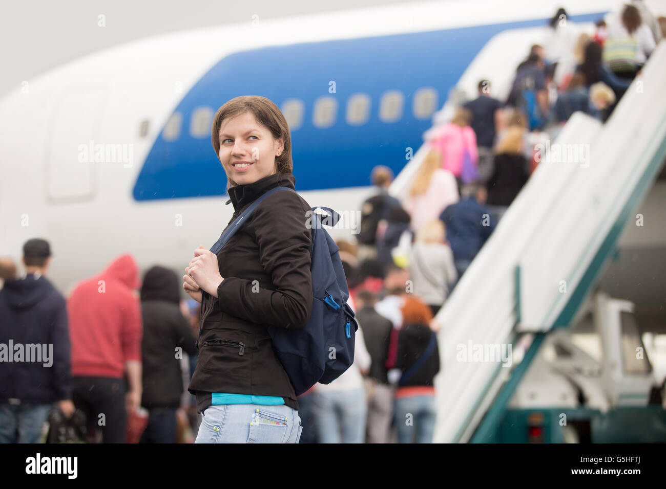 Young Caucasian cheerful smiling woman passenger in 20s travelling with backpack, boarding airplane, people climbing ramp Stock Photo