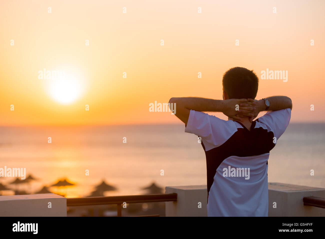 Young man standing on the balcony in carefree pose with hands behind his head, enjoying beautiful colorful sunrise or sunset sea Stock Photo