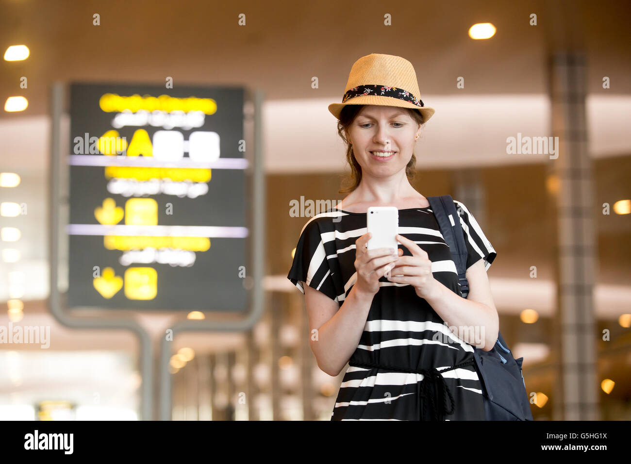 Smiling young woman in straw hat in her 20s walking in modern airport terminal building, holding smartphone, looking at screen Stock Photo