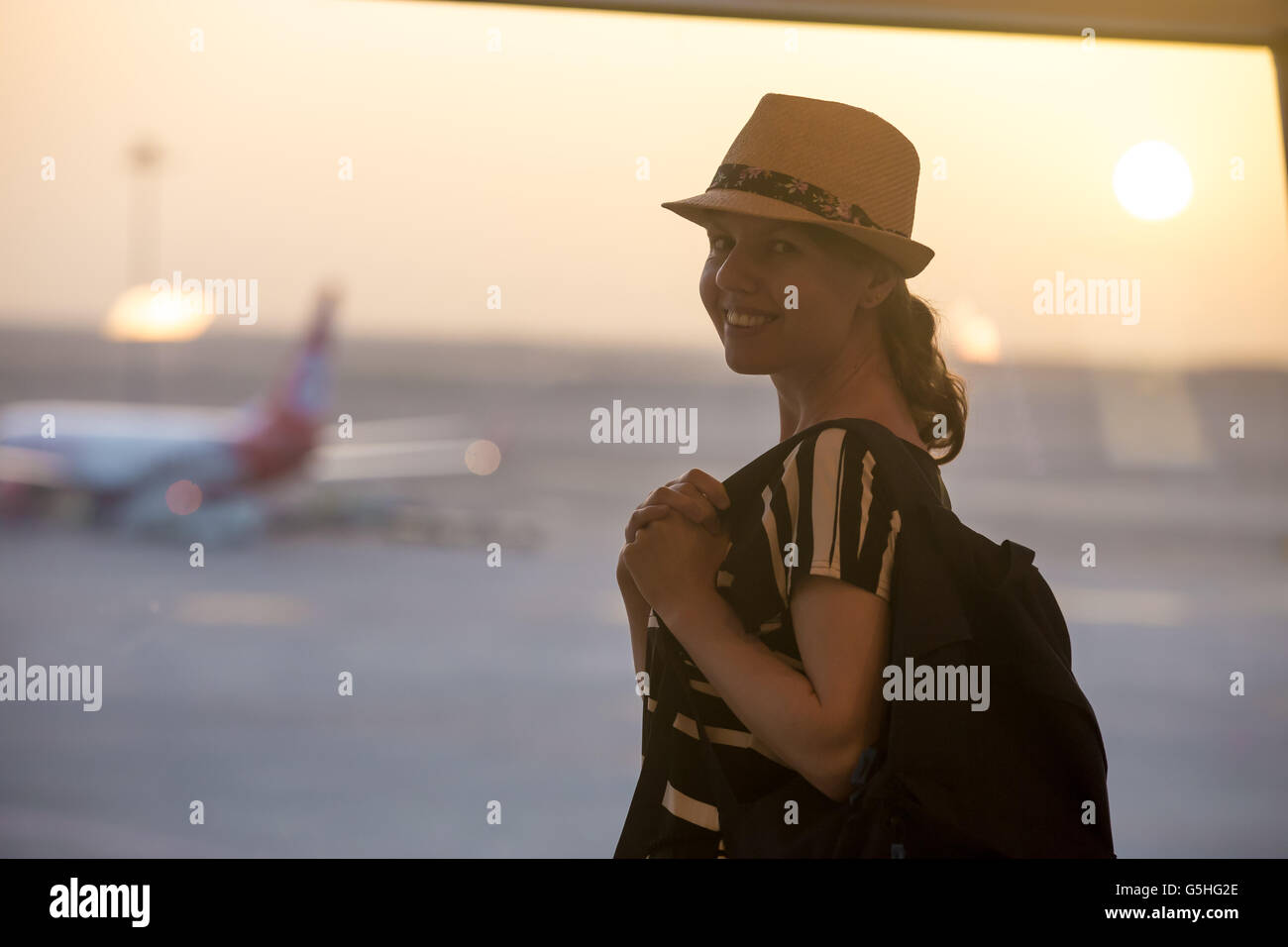Young happy smiling woman in 20s waiting for flight in modern airport terminal building at sunrise or sunset, standing in front Stock Photo
