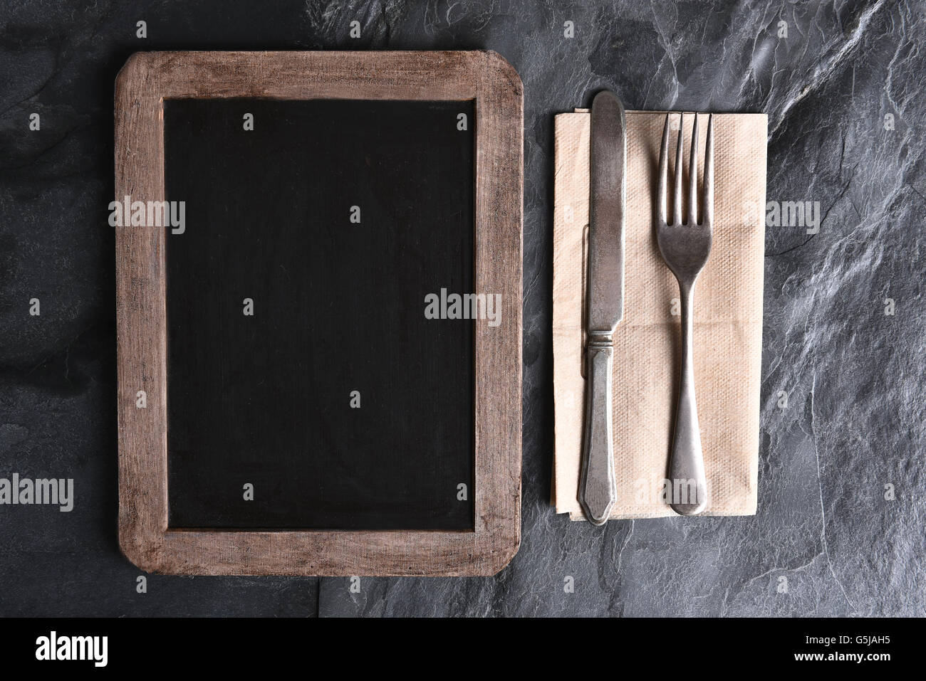 High angle view of a blank chalkboard menu next to silverware on a napkin. Strong side light on a slate table. Stock Photo
