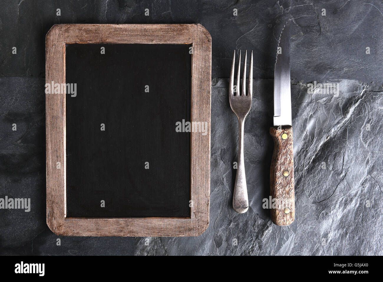 Top view of a knife and fork next to a blank menu chalkboard. Strong side light on a black slate table. Stock Photo