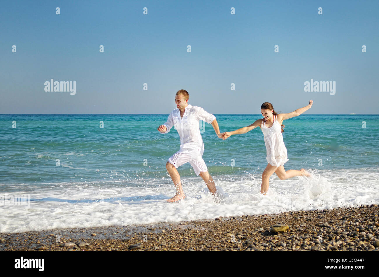 Happy young couple runs having joined hands on the sea coast. The guy and girl dressed in easy white clothes. On faces expressio Stock Photo