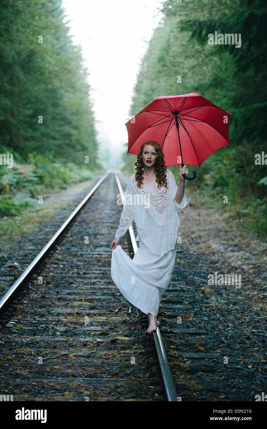 Caucasian woman walking on train tracks with umbrella in forest Stock Photo