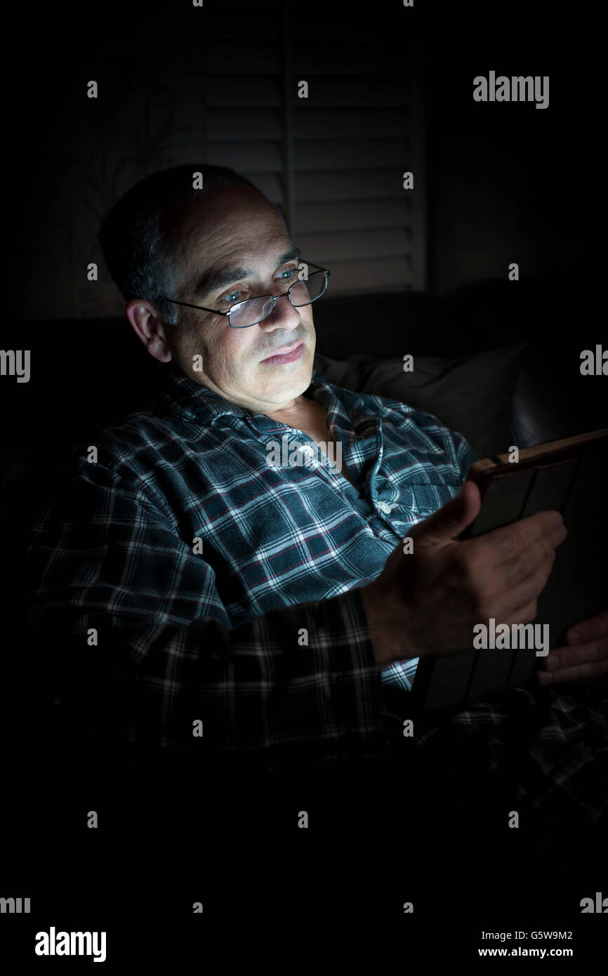 Middle aged man wearing pajamas reading tablet at night in bed with serious expression Stock Photo