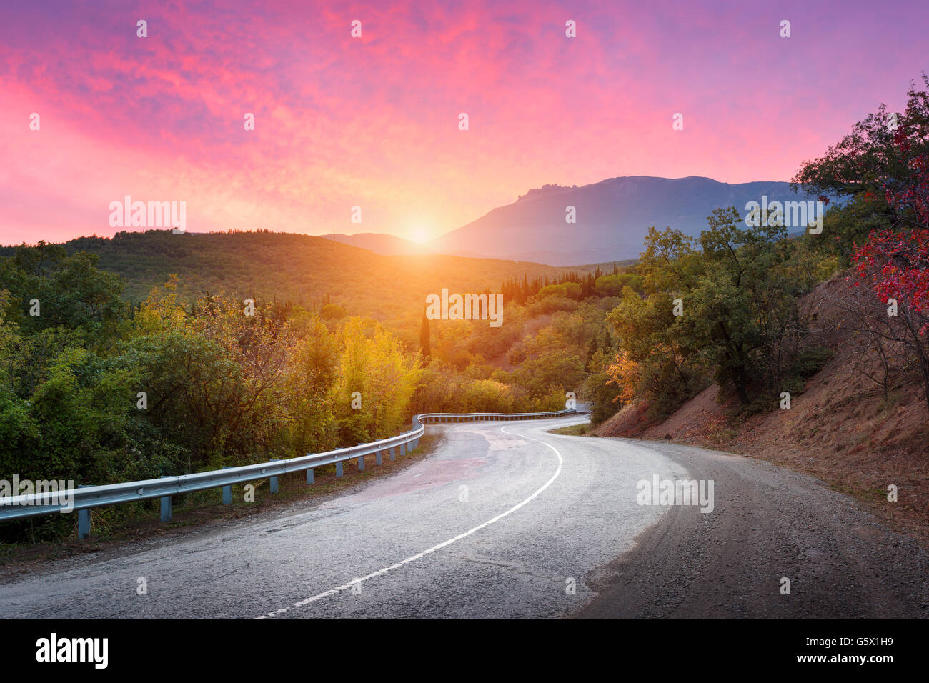 Mountain road passing through the forest with dramatic colorful sky and red clouds at colorful sunset in summer. Mountain landsc Stock Photo