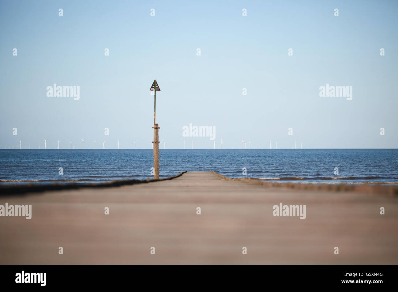 A wooden jetty stretching out into the sea with a wind farm in the distance on the horizon Stock Photo