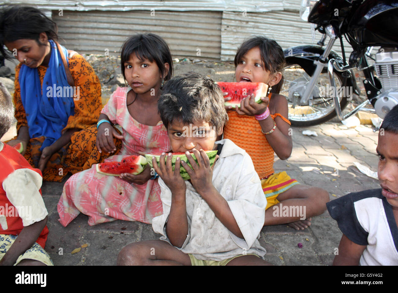 A poor and hungry Indian boy eating a watermelon, his family sitting in