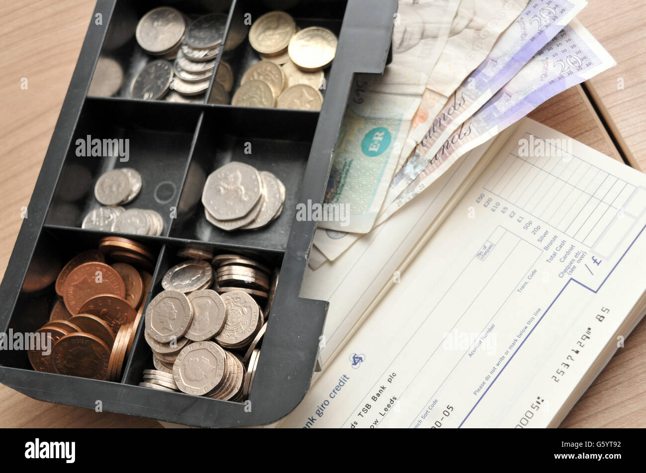 A tray of British coins next to bank notes and a paying in book sat on a backdrop of a wooden work table Stock Photo
