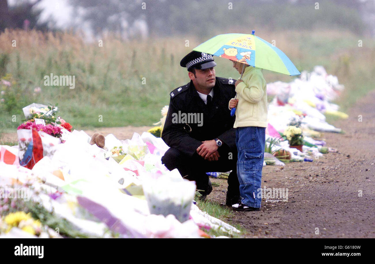 A small girl comes to lay flowers and pay her respects with the help of a police officer beside the rest of the flowers laid at Wangford Road, Lakenheath, Suffolk, near the site where the bodies of Holly Wells and Jessica Chapman were found. Stock Photo