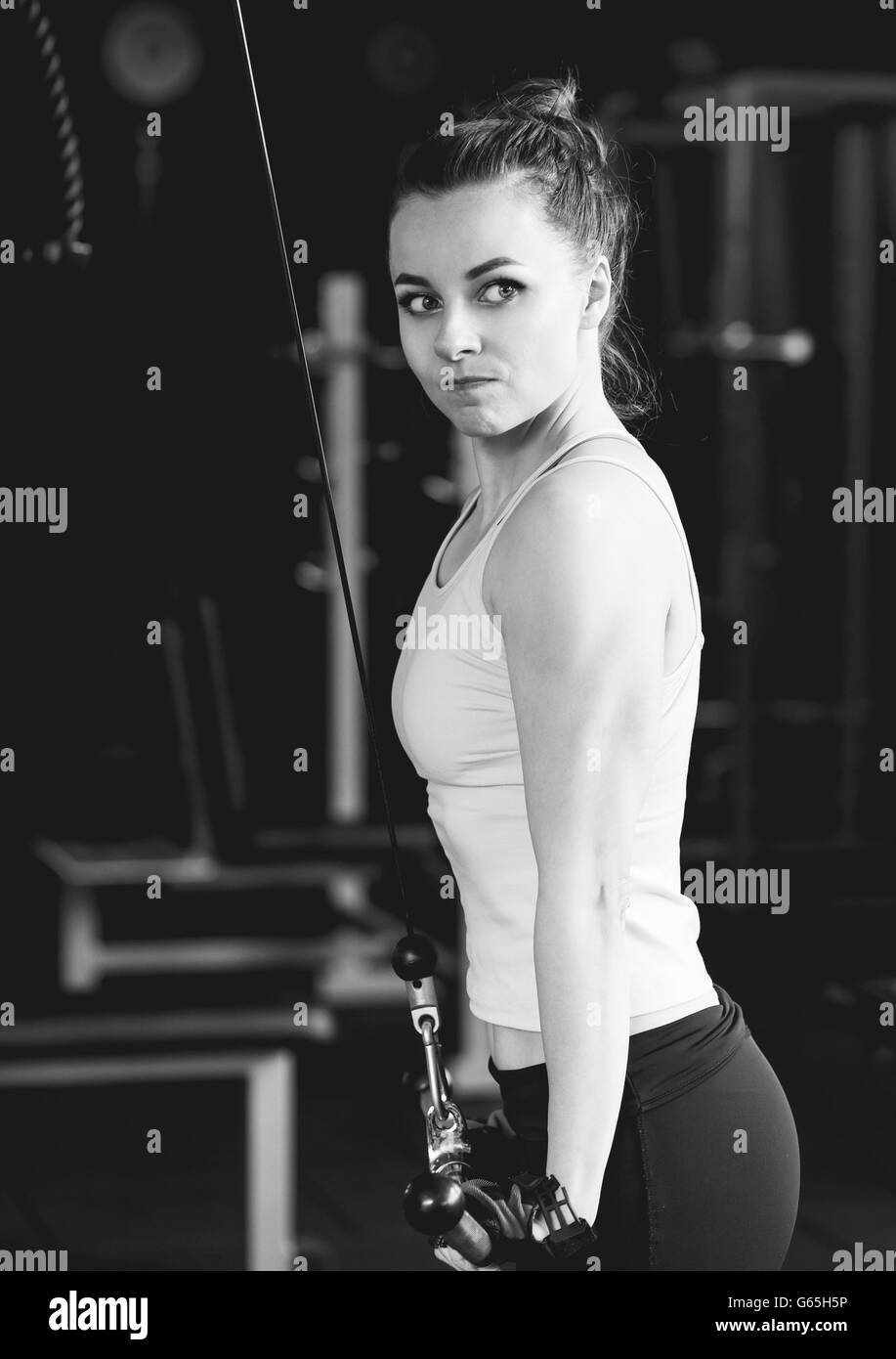 Young slim woman doing pushdown on cable machine in gym. Athletic girl training triceps in fitness center. Black and white image Stock Photo