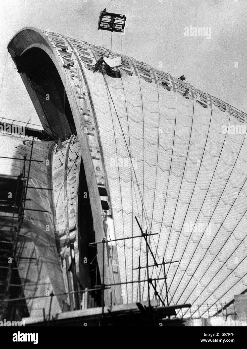 Workmen use a big crane to swing a tile-covered concrete section of the outer casing into place on the Sydney Opera House. More than a million gleaming white tiles will be used before the roof is completed by March but already it presents the striking appearance of soaring white shells reaching out skywards. Construction of the building commenced in March 1959 and proceeded in slow stages over the next fourteen years. Stock Photo