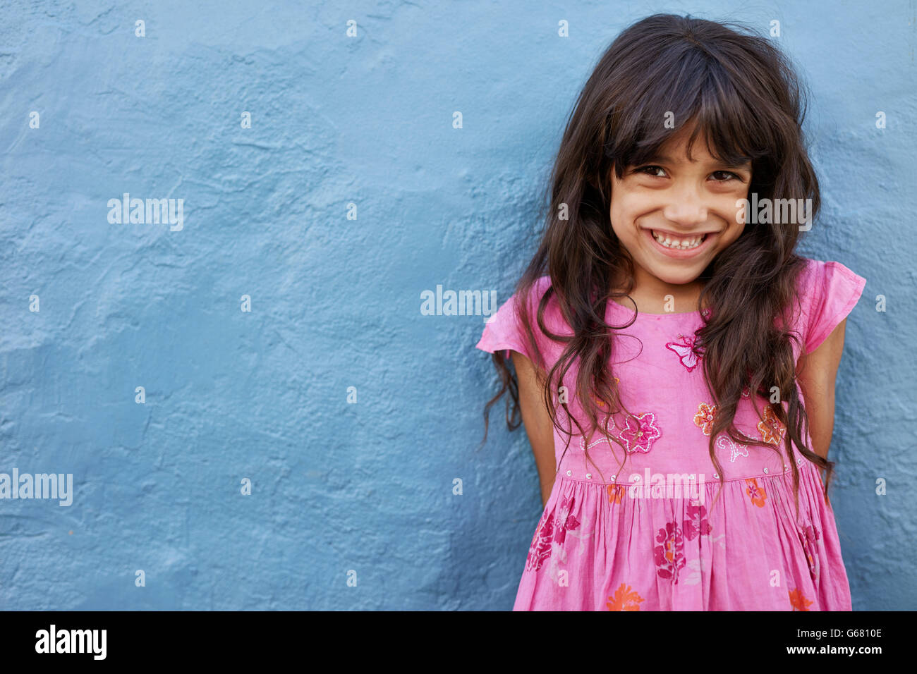 Portrait of cute little girl looking at camera and smiling while standing against blue wall. Innocent young female child with co Stock Photo