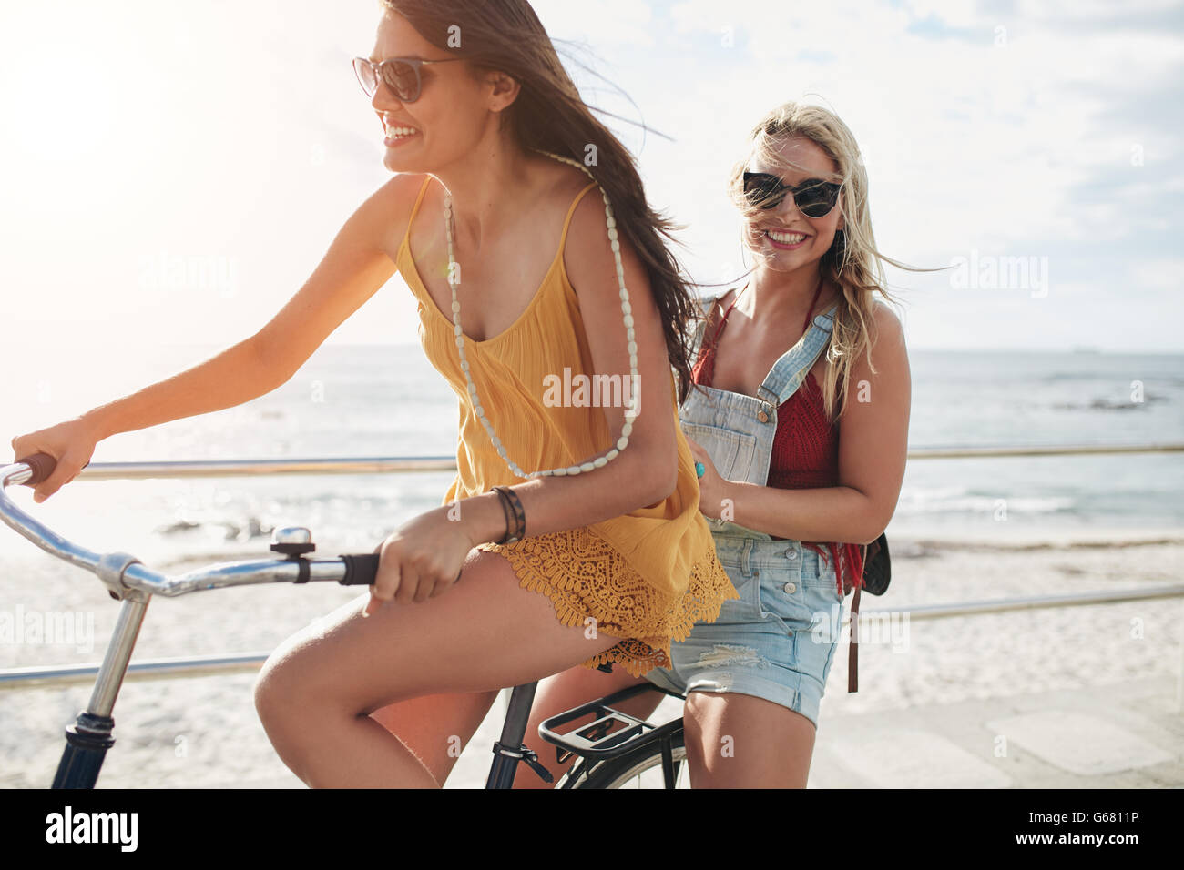 Two stylish young female friends riding together on a bicycle along seaside. Best friends enjoying a day on bike. Stock Photo
