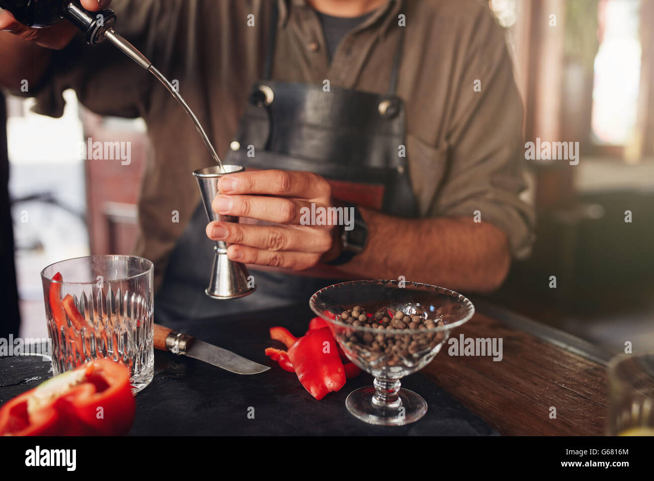 Closeup of bartender hands pouring alcoholic drink into a jigger to prepare a cocktail, with red bell pepper and peppercorn seed Stock Photo