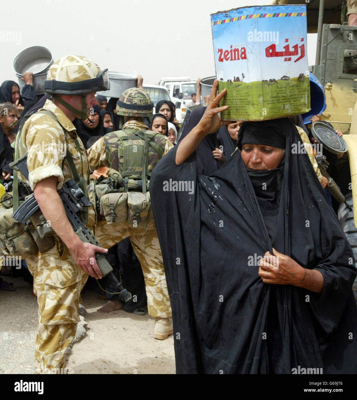 A women carries off water as others wait in a queue with an empty containers which they hope to fill from a tanker, while soldiers from the British 23 Pioneer Regiment watch over the line in the southern Iraqi town of Safwan. * The British forces are helping the local people with humanitarian aid. Stock Photo