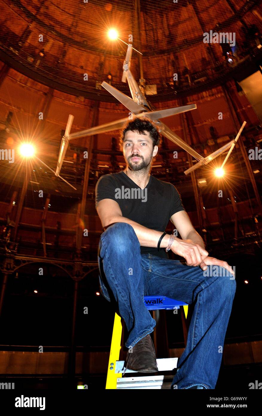 British Artist Conrad Shawcross sits under his artwork titled 'Timepiece', which is three revolving hands with lights at each end to replicate Clock hands, at a press preview at The Rounhouse, in north London, where the modern art will be on display for a month. Stock Photo
