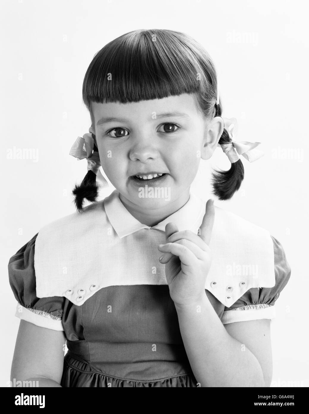 1950s PORTRAIT SMILING GIRL HAIR IN PIGTAILS POINTING FINGER LOOKING AT CAMERA Stock Photo