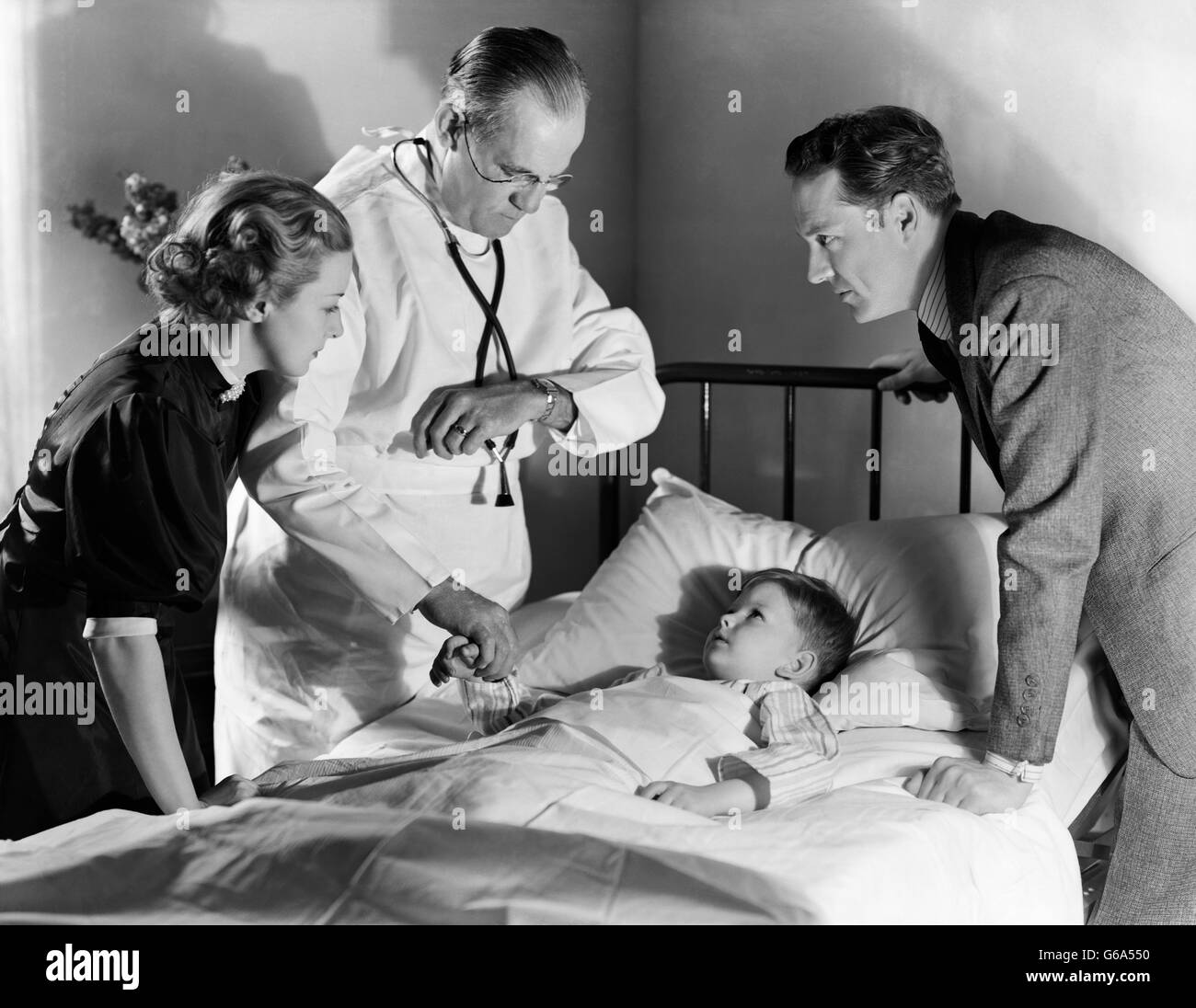 1930s CONCERNED MOTHER FATHER WITH DOCTOR CHECKING PULSE SMALL BOY IN HOSPITAL BED Stock Photo